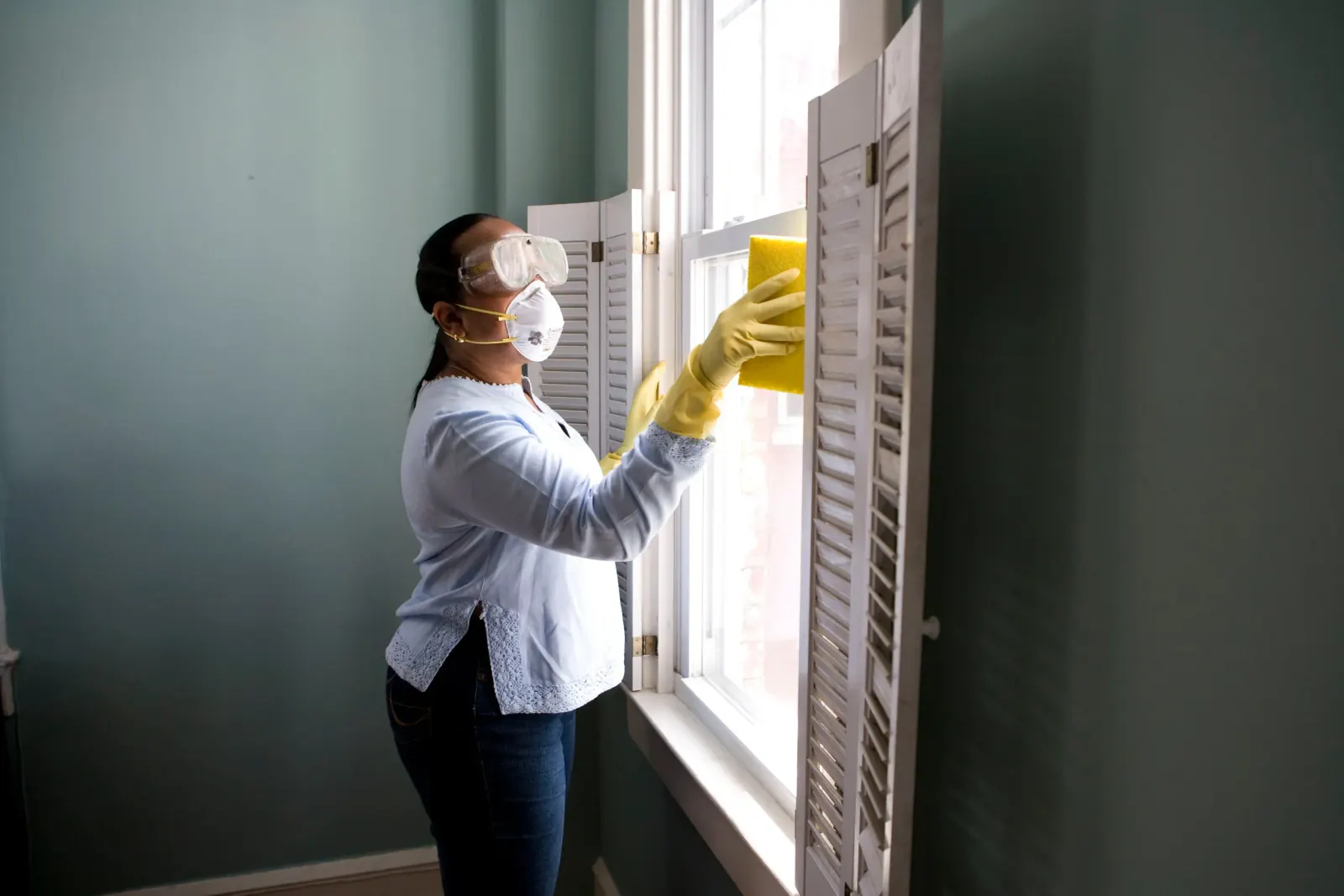 Cleaner in gloves and protective eyewear wiping a window frame and shutters